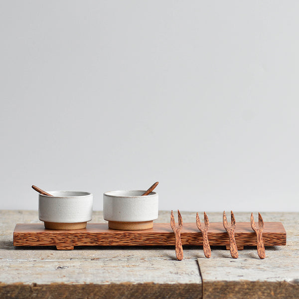 Two white stoneware bowls with wooden spoons on a coconut wood tray, with four small wooden forks beside them.