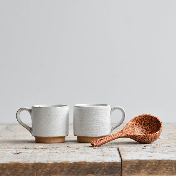 Two stackable snow white stoneware espresso cups with unglazed foot rings next to a hand-carved coconut palm wood scoop. placed in a wooden table. Nom Living