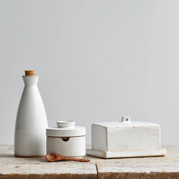 A coordinating snow white ceramic kitchen set with a stoneware oil bottle, a covered butter dish, and a matching lidded sugar bowl with a spoon. Nom Living