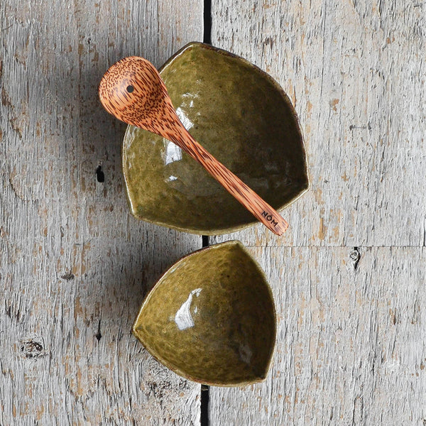 Handmade deep green triangular ceramic olive serving bowl next to a smaller pip dish and a coconut wood draining spoon. Shot from above. Nom Living