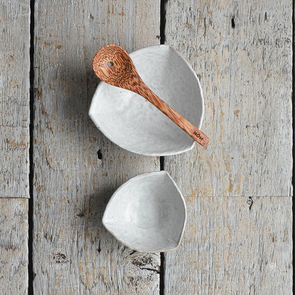Handmade snow white triangular ceramic olive serving bowl next to a smaller pip dish and a coconut wood draining spoon. Shot from above. Nom Living
