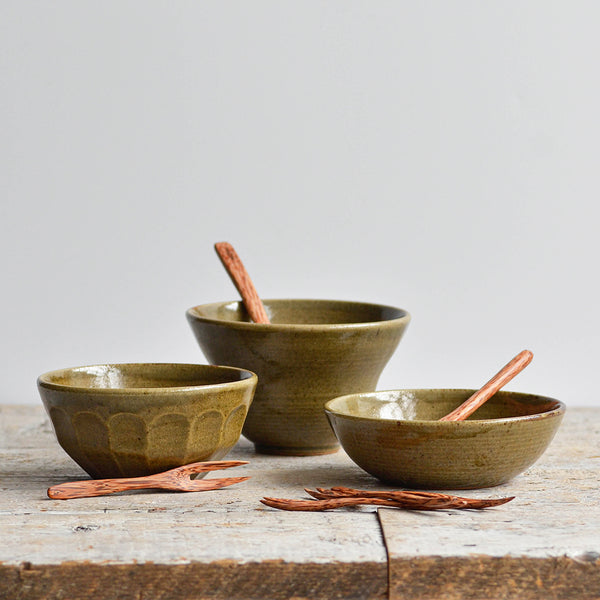 Nom Living - Set of three handcrafted green stoneware snack bowls with coconut wood spoons and cocktail forks on a rustic wooden table.