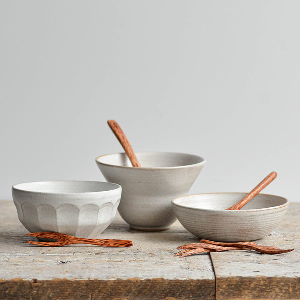 Nom Living - Set of three handcrafted snow-white stoneware snack bowls with coconut wood spoons and cocktail forks on a rustic wooden table.