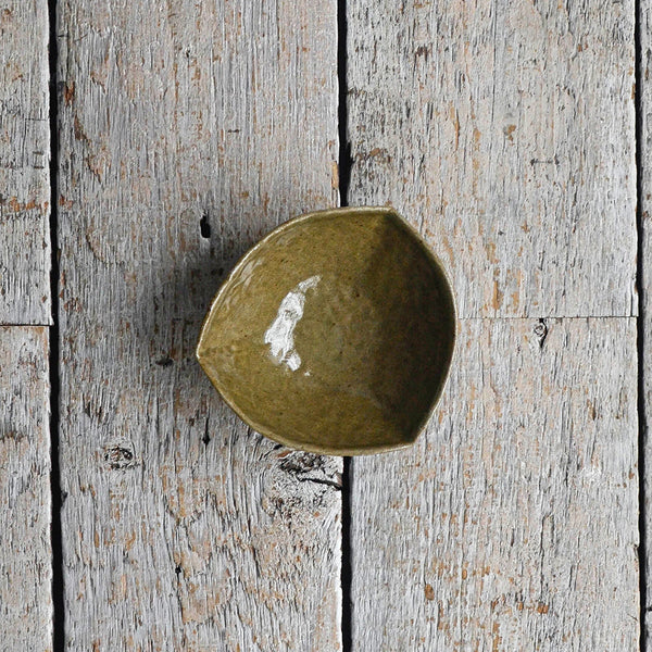 Hand-moulded dark green stoneware bowl in an organic triangular shape with a textured, earthy glaze, designed for serving olives, dips, and small appetizers. placed in a wood table