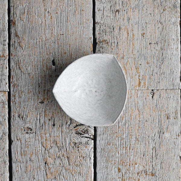 Hand-moulded Snow-White stoneware bowl in a soft, irregular triangular shape with a textured surface, ideal for serving olives, dips, and side dishes placed on a wood table.