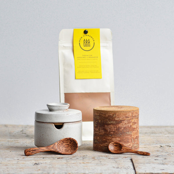 Nom Living - Stoneware white ceramic sugar bowl with wooden spoon, brown spice container, and pouch of premium ground cinnamon on a wooden table. 