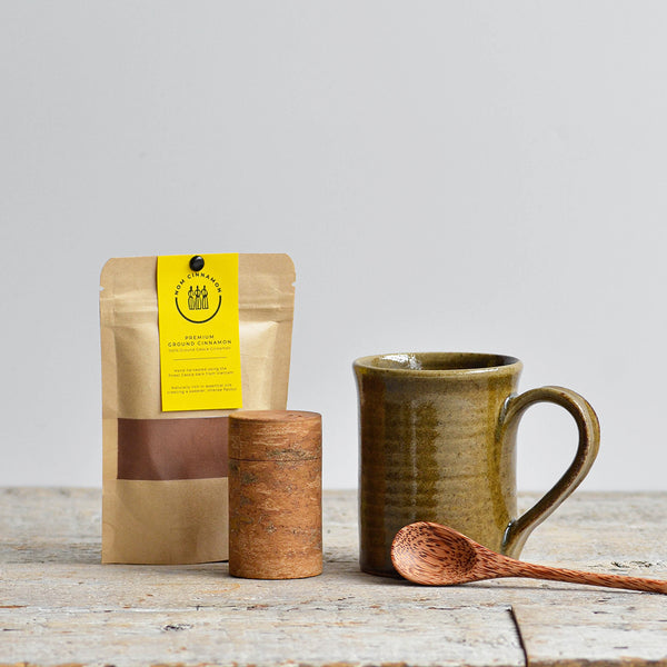 Rustic green ceramic mug with wooden spoon, brown spice container, and pouch of premium ground cinnamon on a wooden table. Nom Living