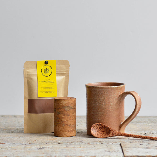 Stoneware brown ceramic mug with wooden spoon, brown spice container, and pouch of premium ground cinnamon on a wooden table. Nom Living